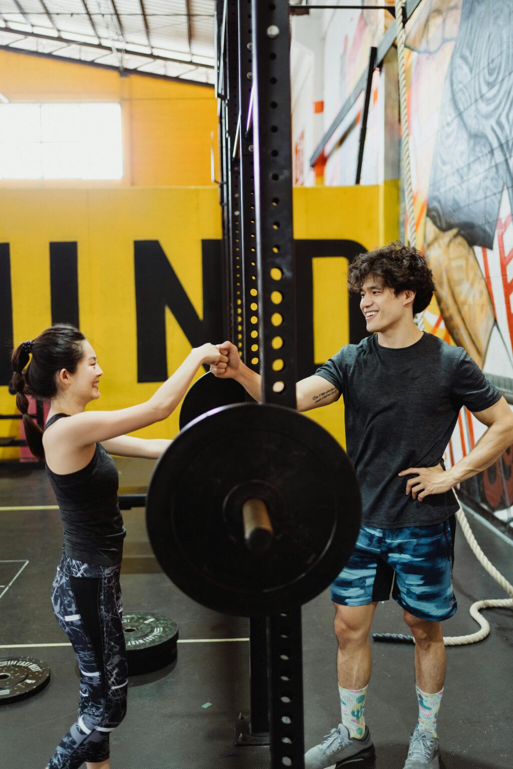 A man and a woman share a fist bump in an energetic gym setting, surrounded by fitness equipment.